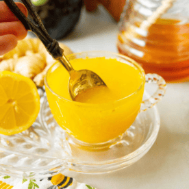 A golden spoon is dipped into a glass of bright yellow liquid, surrounded by a sliced lemon, ginger, and a jar of honey on a patterned cloth. This scene captures the essence of an immunity tea bombs recipe in its vibrant detail.