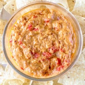 A glass bowl brimming with creamy, spicy smoked chorizo queso, interspersed with diced tomatoes, surrounded by tortilla chips on a platter.