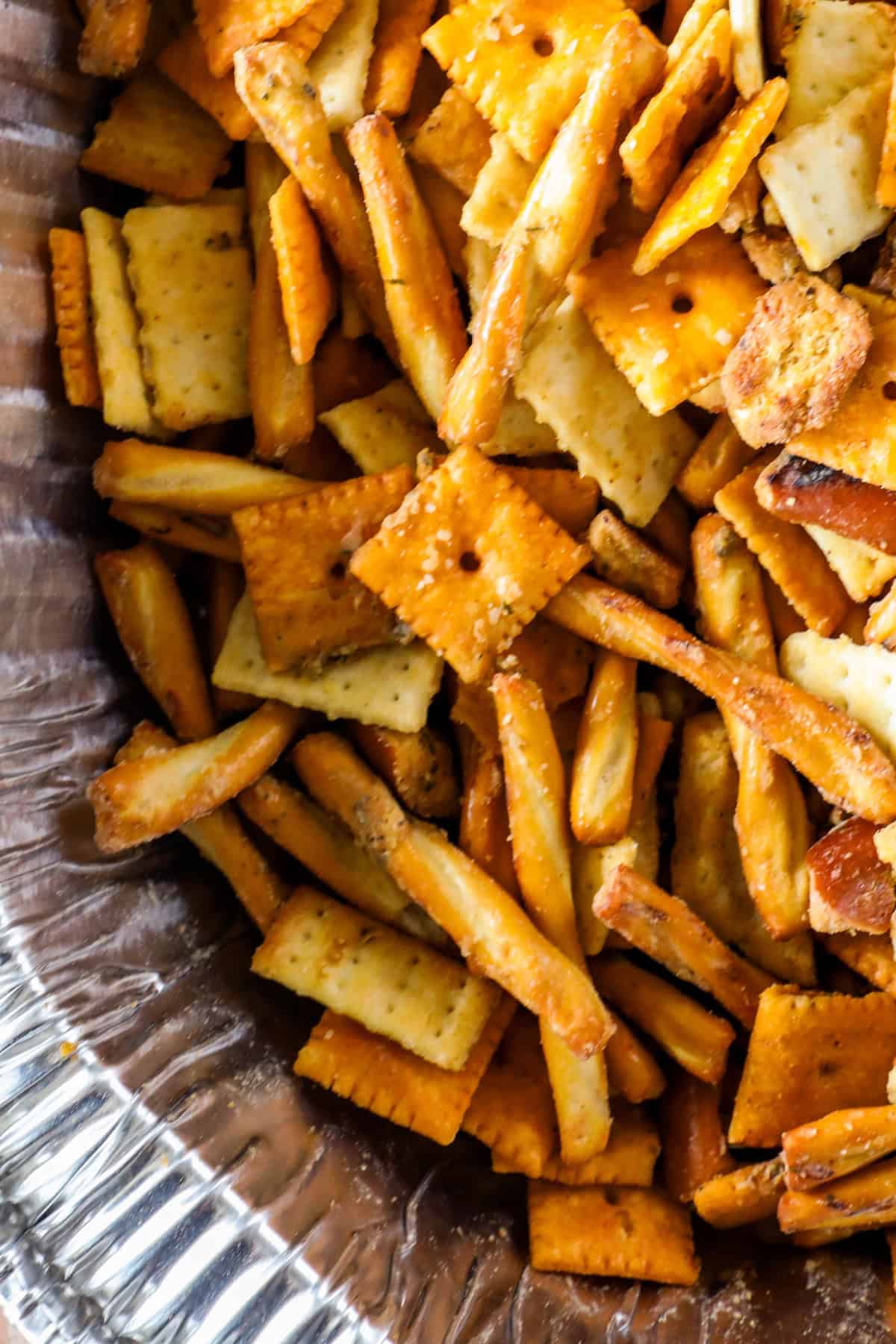 A close-up of a smoked trail mix featuring a blend of square crackers and pretzel sticks nestled in a foil-lined container.