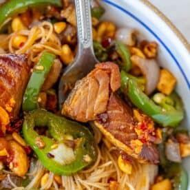 Close-up of a plate with noodles, sliced beef, jalapeños, chopped nuts, and onions, garnished with red pepper flakes. A fork is resting on the plate.