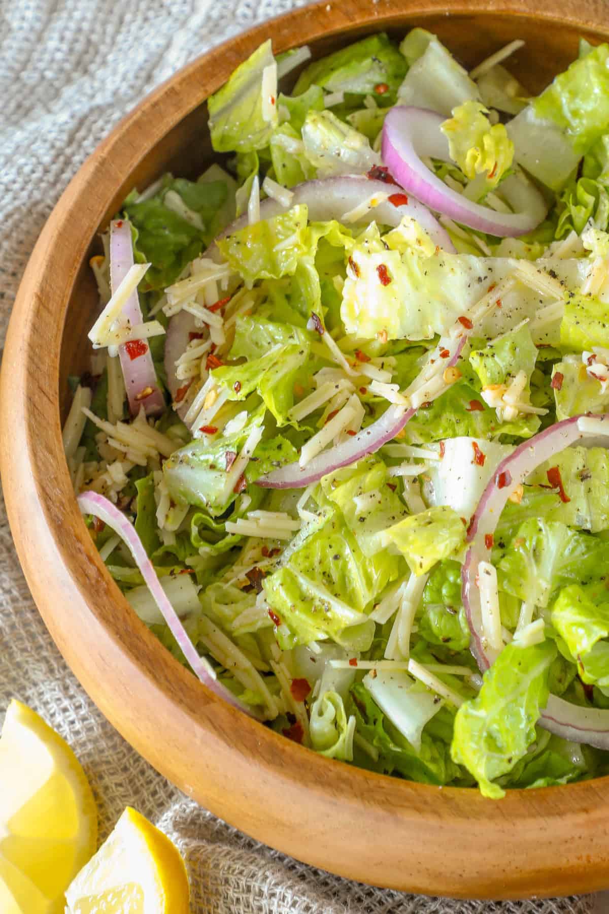 A wooden bowl filled with a romaine parmesan salad features crisp lettuce, red onion slices, cheese, and a sprinkle of red pepper flakes. Lemon wedges rest nearby on a textured fabric surface.