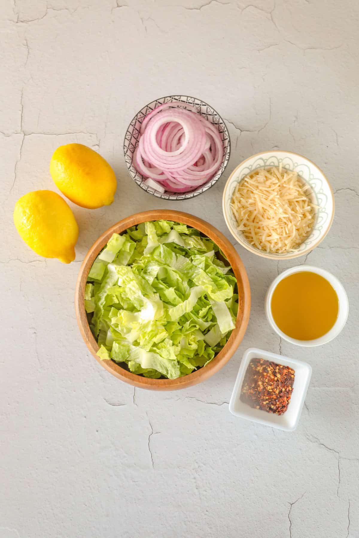 Ingredients for a romaine salad, featuring a wooden bowl of chopped lettuce, two lemons, sliced red onions, grated Parmesan cheese, a small bowl of oil, and a dish of red pepper flakes.