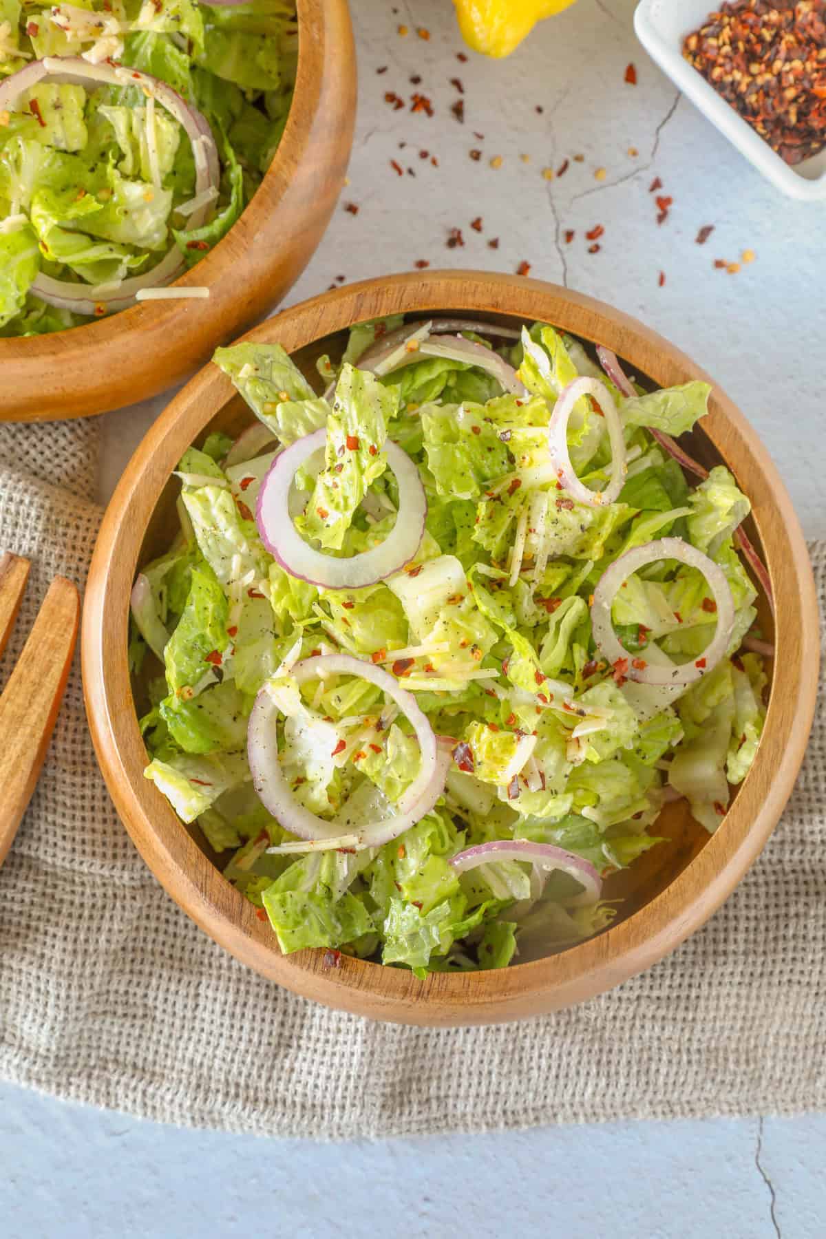 A wooden bowl brimming with a romaine salad, featuring fresh lettuce and onion rings, sprinkled with parmesan on a light surface. A cloth napkin and a partial view of another bowl complete this inviting scene.