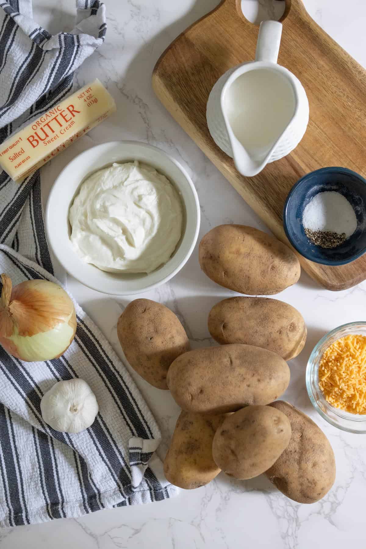 Ingredients for making cheesy creamy mashed potatoes include russet potatoes, butter, cream, shredded cheese, onion, garlic, salt, and pepper. They’re beautifully displayed on a striped towel and wooden board.
