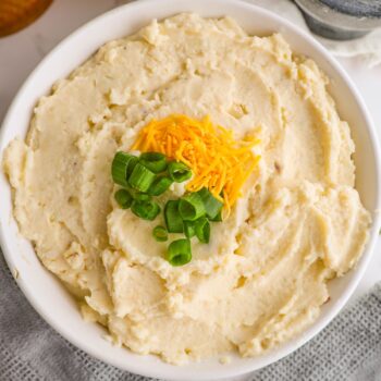 A bowl of cheesy, creamy mashed potatoes topped with shredded cheese and chopped green onions.