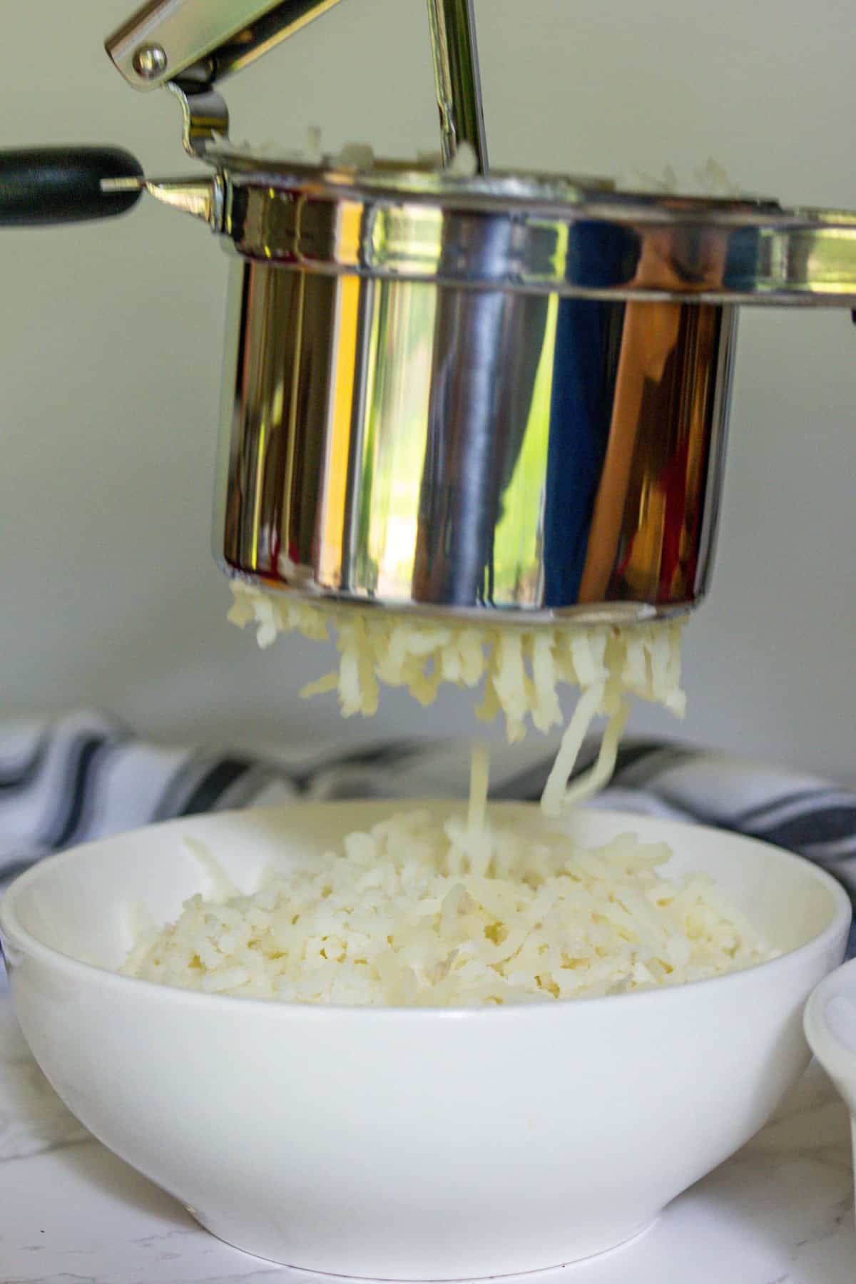 A potato ricer presses cooked potatoes into thin strands over a white bowl, creating the perfect base for cheesy creamy mashed potatoes.
