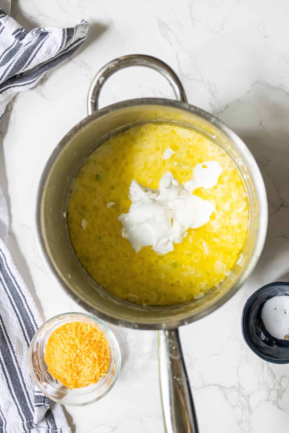 A pot of cheesy corn chowder topped with cream sits on a marble surface, accompanied by a bowl of grated cheese and a dish of salt. Nearby, a striped cloth adds charm to the scene.