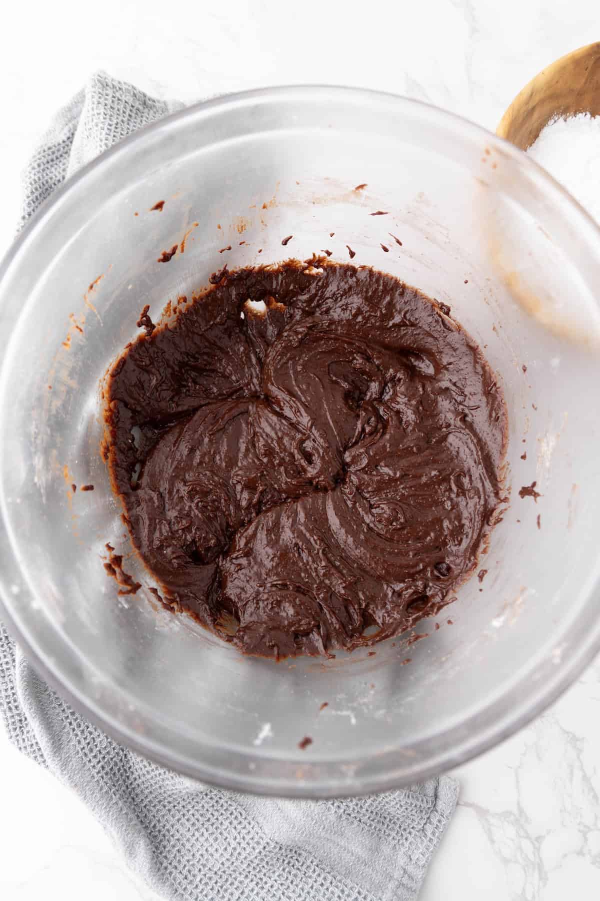 A glass bowl holds chocolate brownie batter atop a gray napkin, paired perfectly with a wooden bowl of salt nearby, setting the stage for delightful chocolate crinkle cookies.