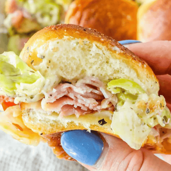 Close-up of a hand holding a classic deli slider with ham, lettuce, and sauce in a soft bun. The person's nails are painted blue.