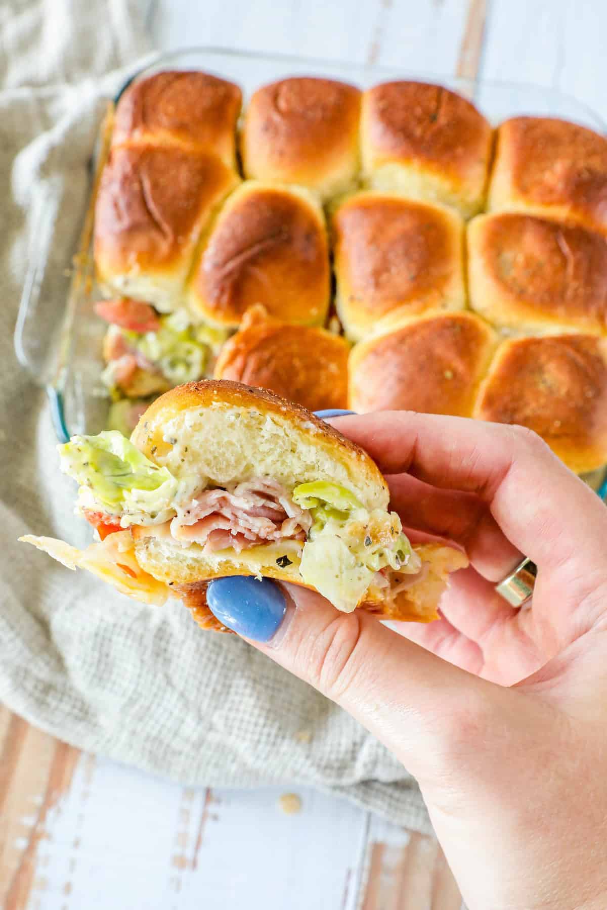 A hand holds a classic deli slider filled with lettuce and meat. In the background, a baking dish brimming with these delightful sliders rests on a cloth.