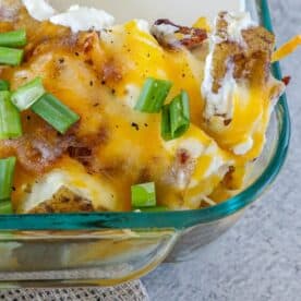 Close-up of a loaded baked potato casserole featuring melted cheese, crispy bacon, and fresh green onions in a glass dish.