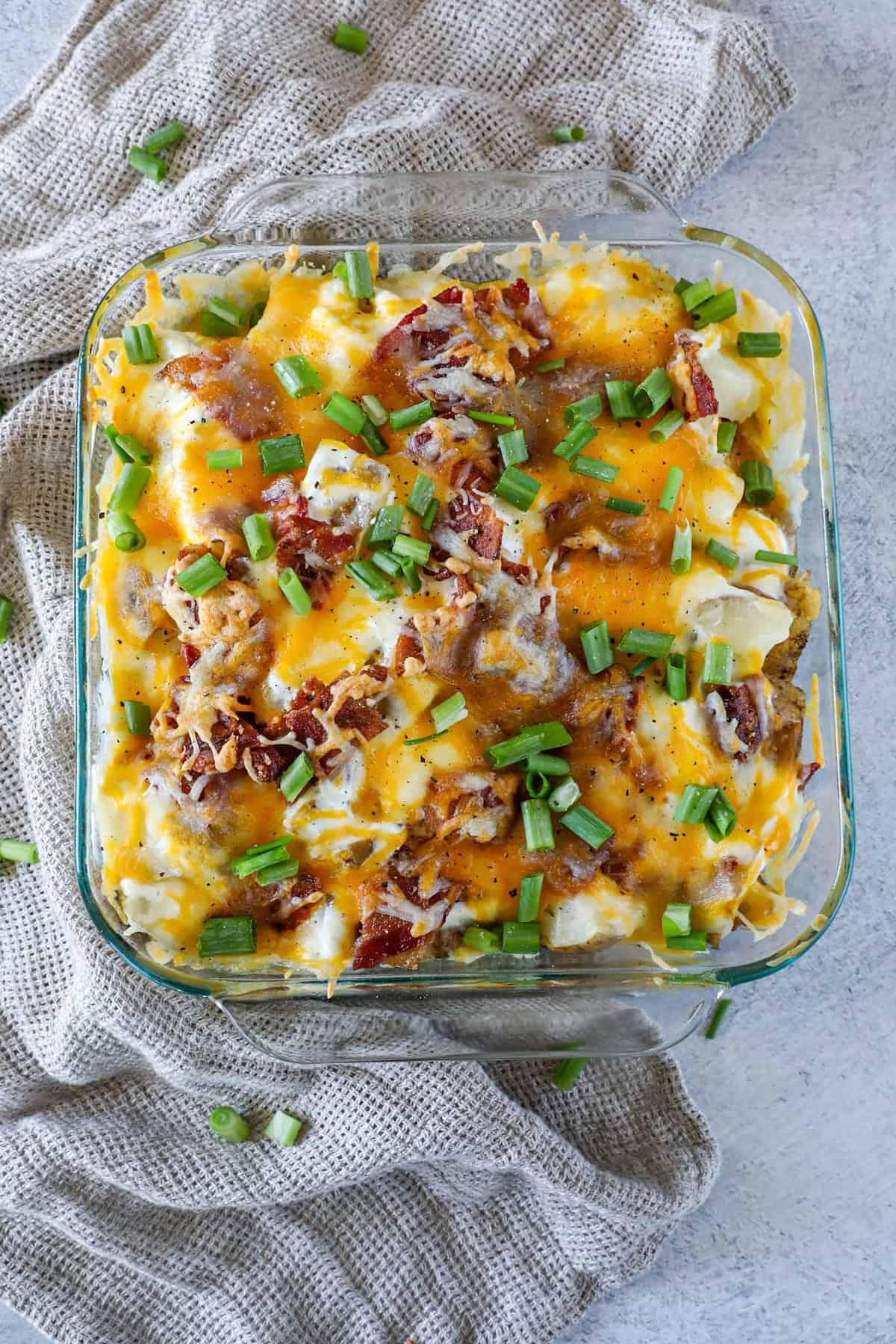 Loaded baked potato casserole topped with melted cheese, bacon bits, and chopped green onions in a glass baking dish on a textured cloth.