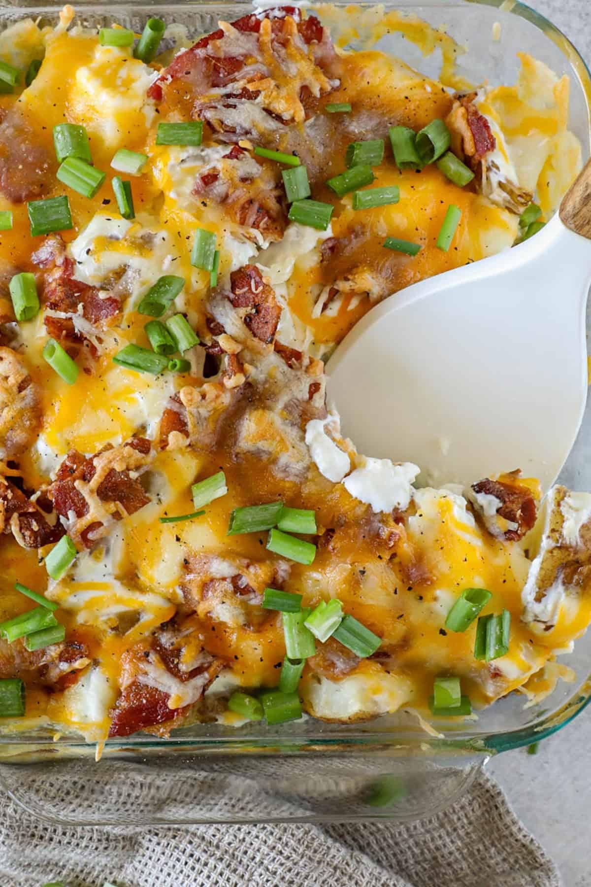 A casserole dish brimming with a loaded baked potato casserole, featuring cheesy goodness and topped with crisp bacon pieces and fresh green onions. A serving spoon eagerly scoops out the deliciousness.