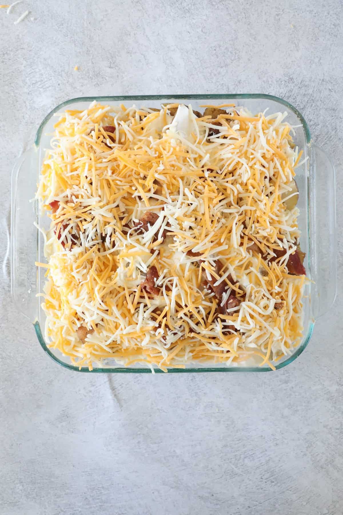 Ultimate Loaded Potato Bake 9 A glass baking dish showcases a loaded baked potato casserole, layered and topped with shredded cheese and crispy bacon bits on a gray surface.