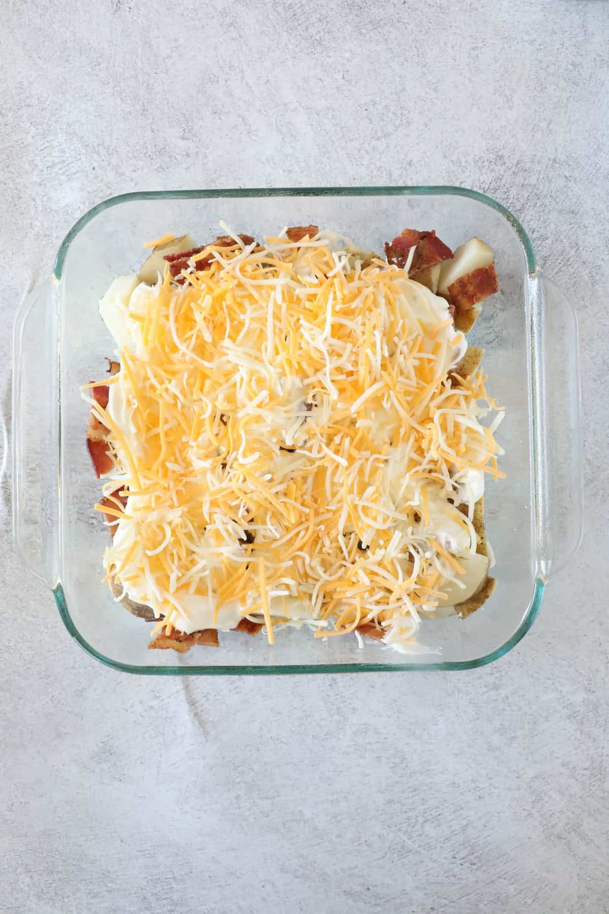 Ultimate Loaded Potato Bake 8 A glass dish filled with layered ingredients, showcasing the essence of a loaded baked potato casserole, with shredded cheese and sliced potatoes, rests on a gray textured surface.