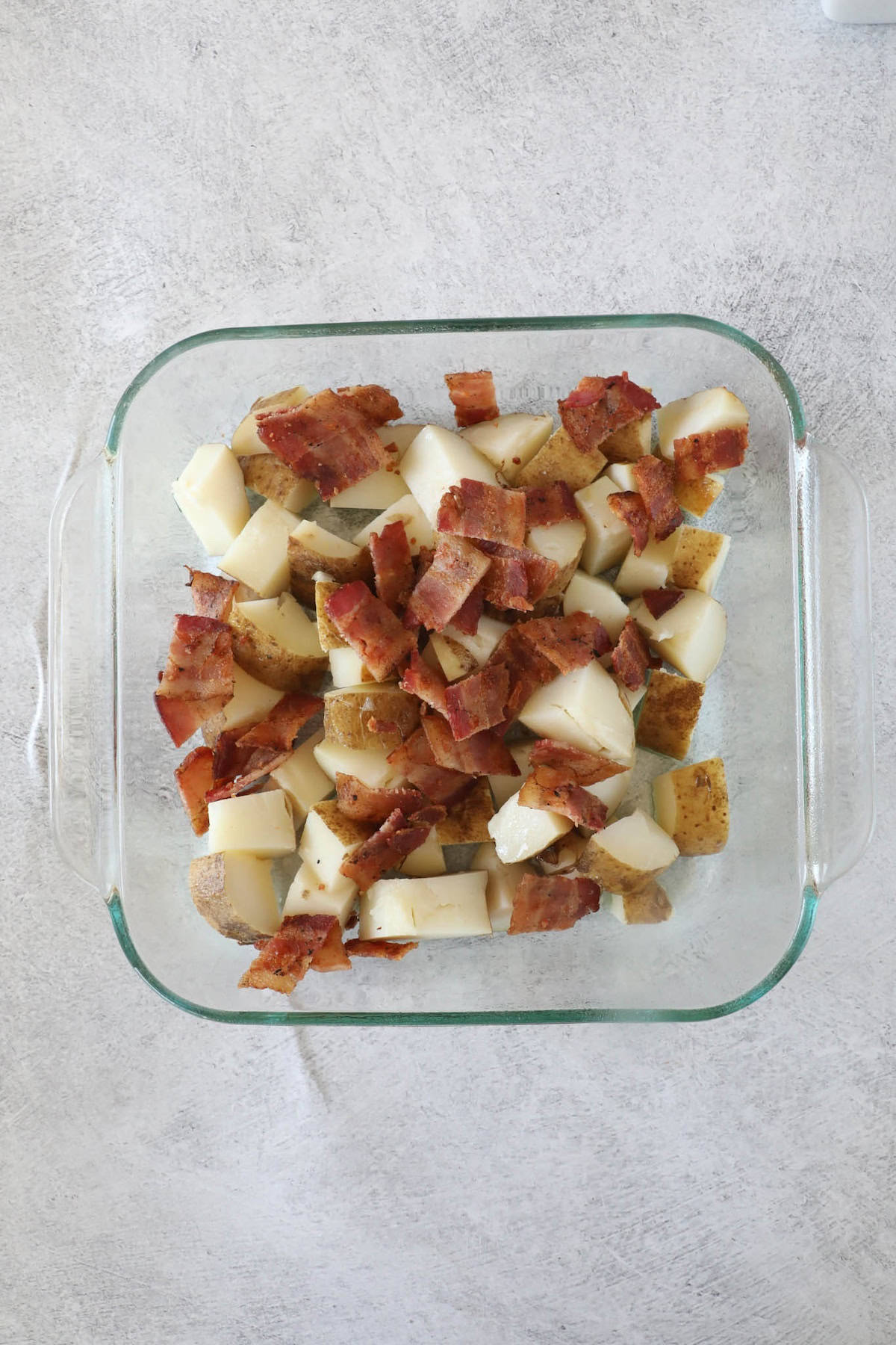 Ultimate Loaded Potato Bake 7 Glass dish with a loaded baked potato casserole, featuring diced potatoes and crumbled bacon atop a light surface.