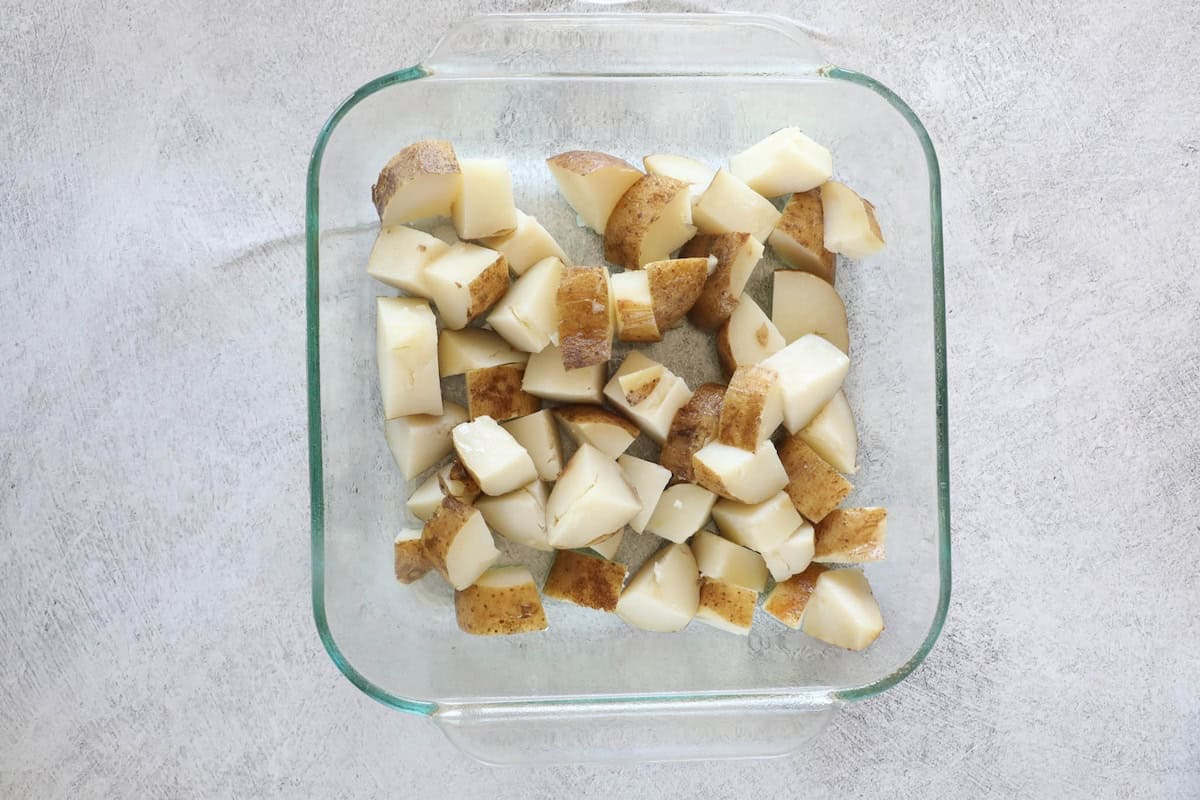 Ultimate Loaded Potato Bake 6 Diced potatoes with skins on rest in a square glass baking dish, ready to transform into a delectable loaded baked potato casserole, set against a textured white surface.
