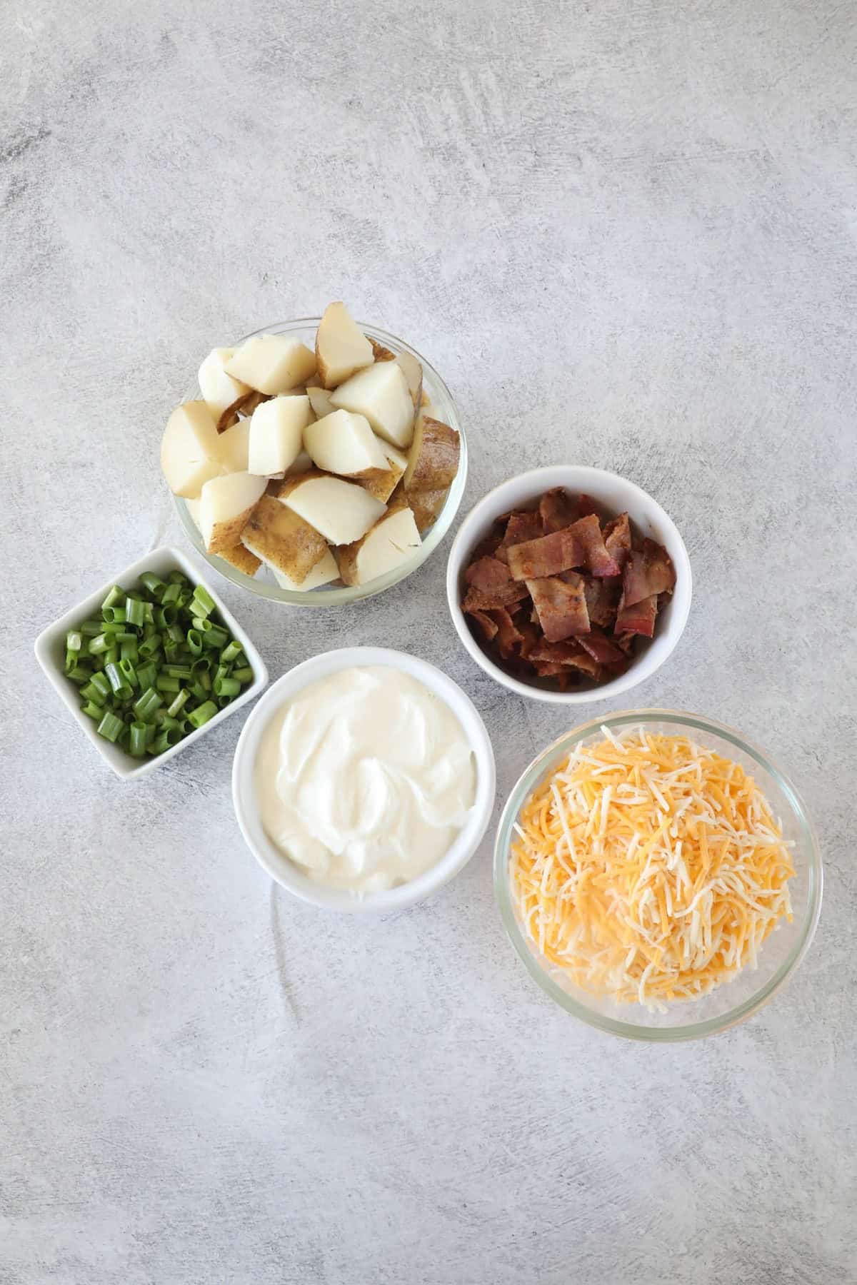 Ultimate Loaded Potato Bake 5 Ingredients for a loaded baked potato casserole: diced potatoes, chopped green onions, crispy bacon pieces, sour cream, and shredded cheese arranged in separate bowls on a gray surface.