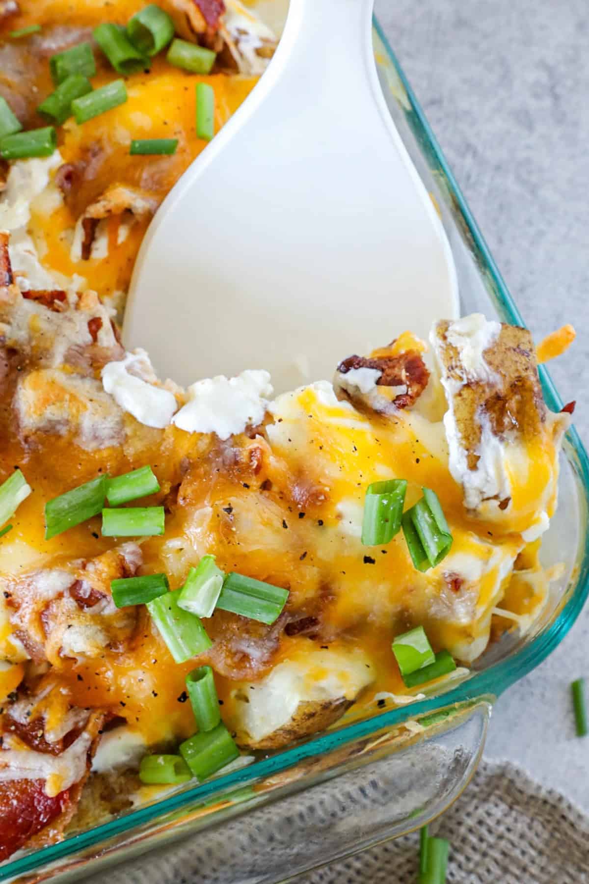 A close-up of a loaded baked potato casserole with melted cheese, bacon, and scallions. A white spoon is scooping a portion from the dish in a clear glass baking pan.