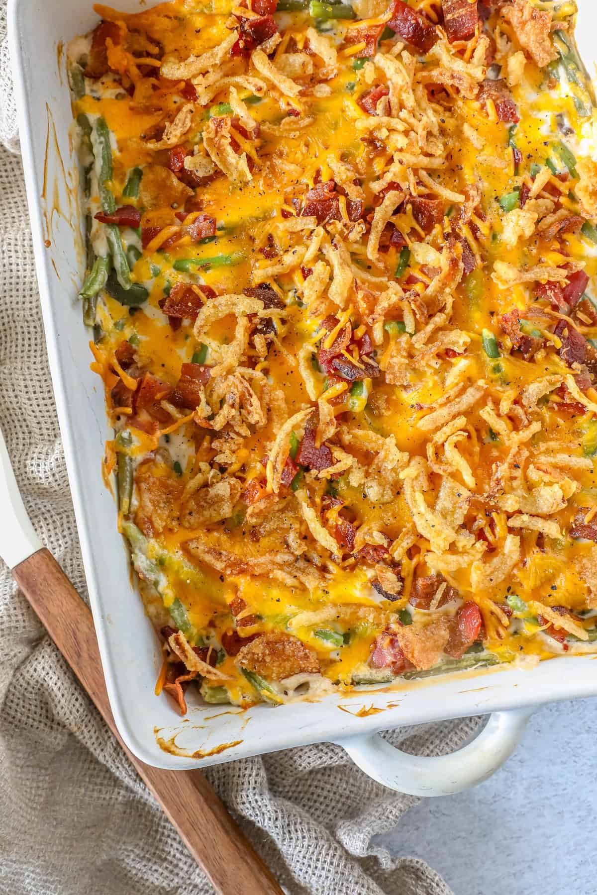 A green bean casserole baked to perfection, featuring melted cheese, crispy onions, and tender green beans, all nestled in a rectangular dish on a textured cloth.