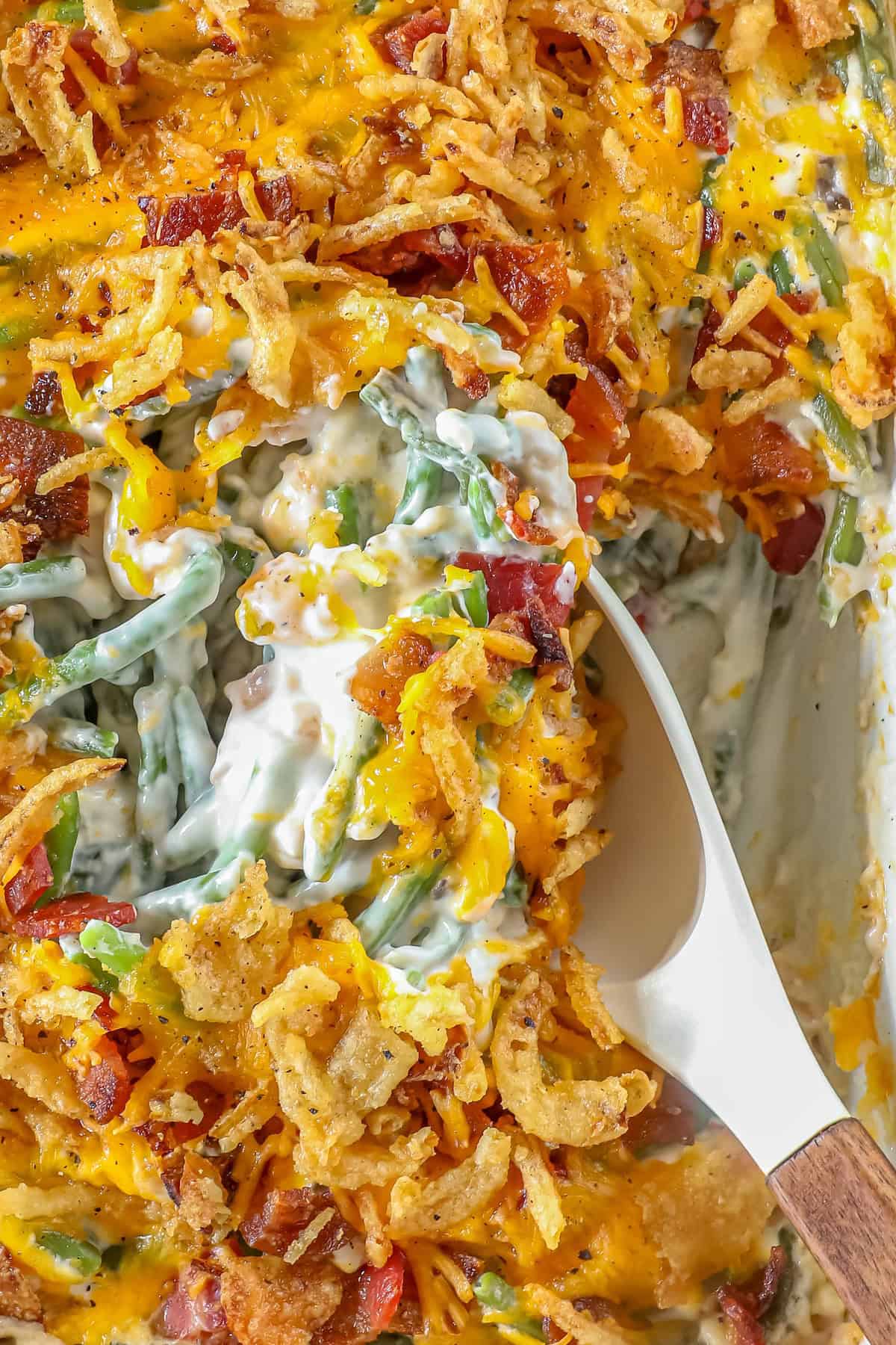 A spoon serving a portion of green bean casserole reveals a close-up of the dish, topped with melted cheese, crispy fried onions, and savory bacon bits.