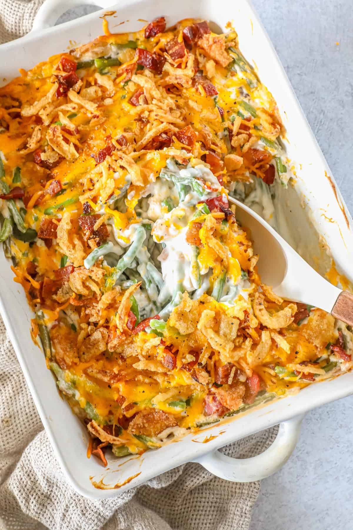 A baked casserole dish with savory green bean casserole topped with melted cheese and crispy onions, a serving spoon partially lifting a delicious portion.