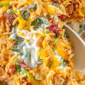 A close-up of a cheesy green bean casserole showcases crispy fried onions and savory bacon pieces atop a creamy sauce, with a spoon ready to serve.