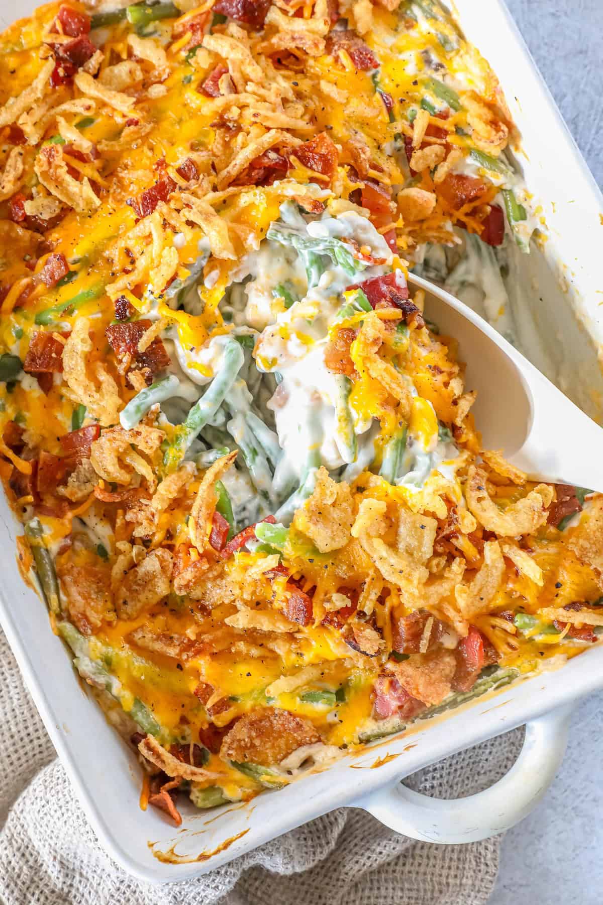 A casserole dish features a classic green bean casserole topped with melted cheese, crispy fried onions, and bacon, while a white serving spoon elegantly scoops a generous portion.