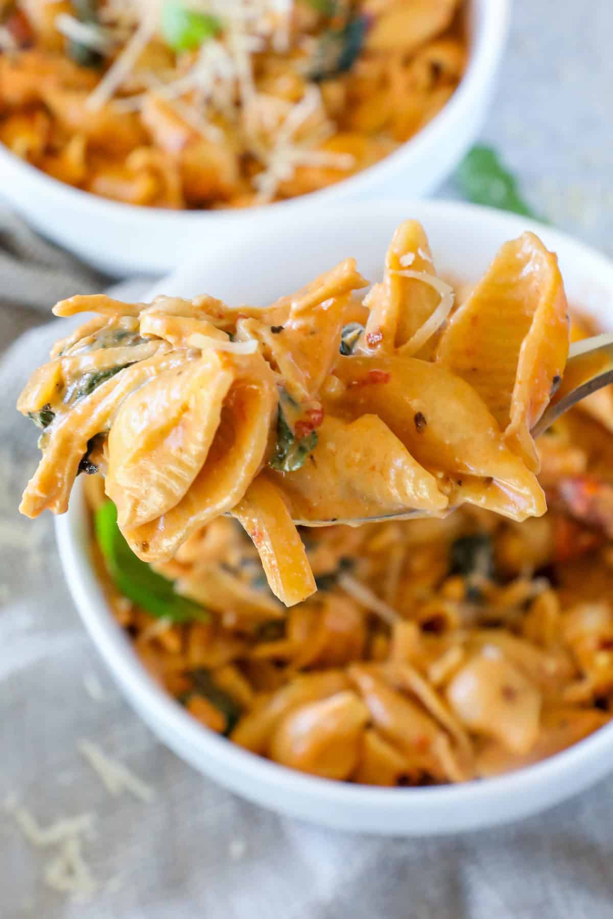 Close-up of a spoon holding creamy pasta shells with spinach and tomatoes, over a bowl of the same dish.