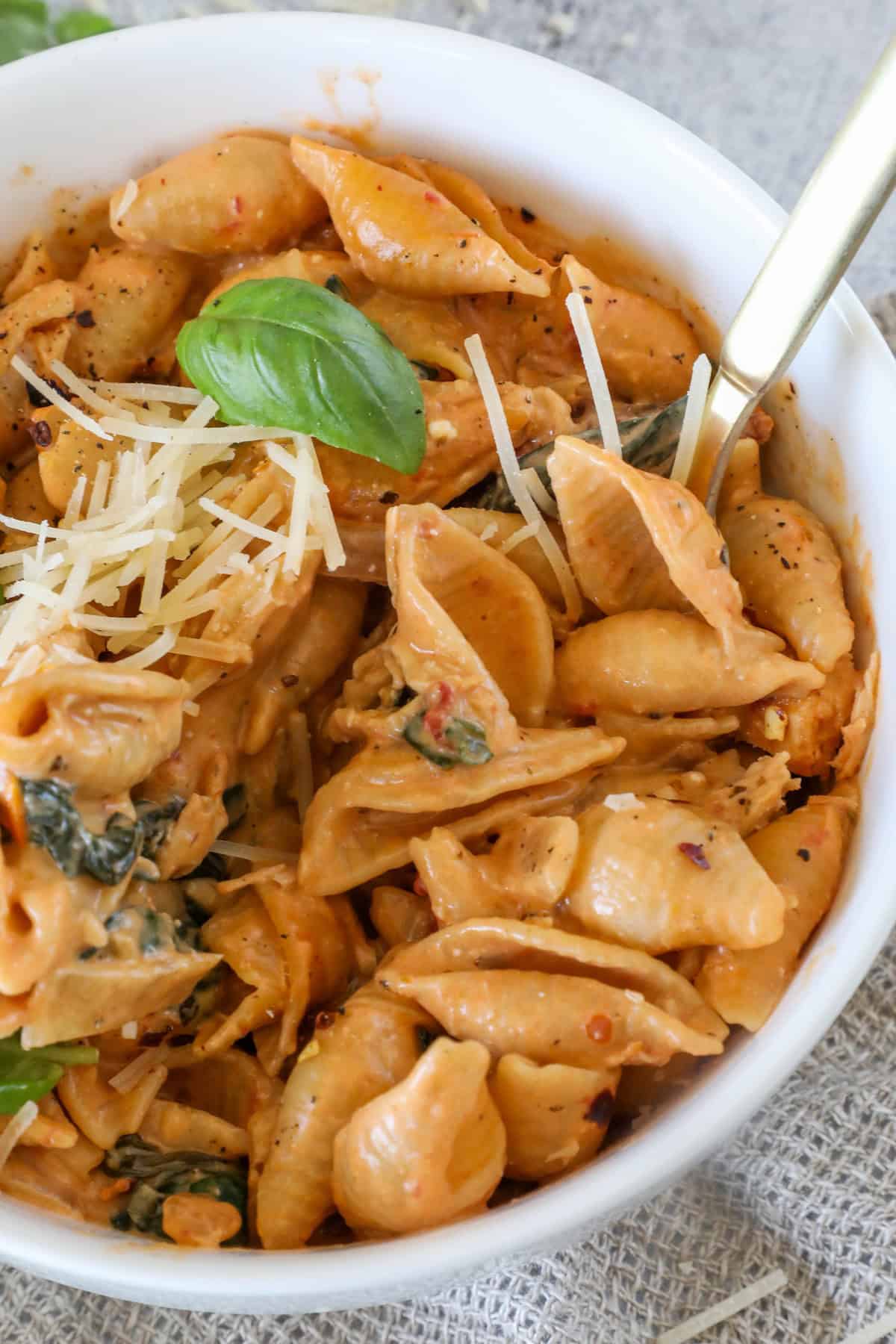 A bowl of creamy pasta shells with spinach, topped with grated cheese and a basil leaf, accompanied by a fork.