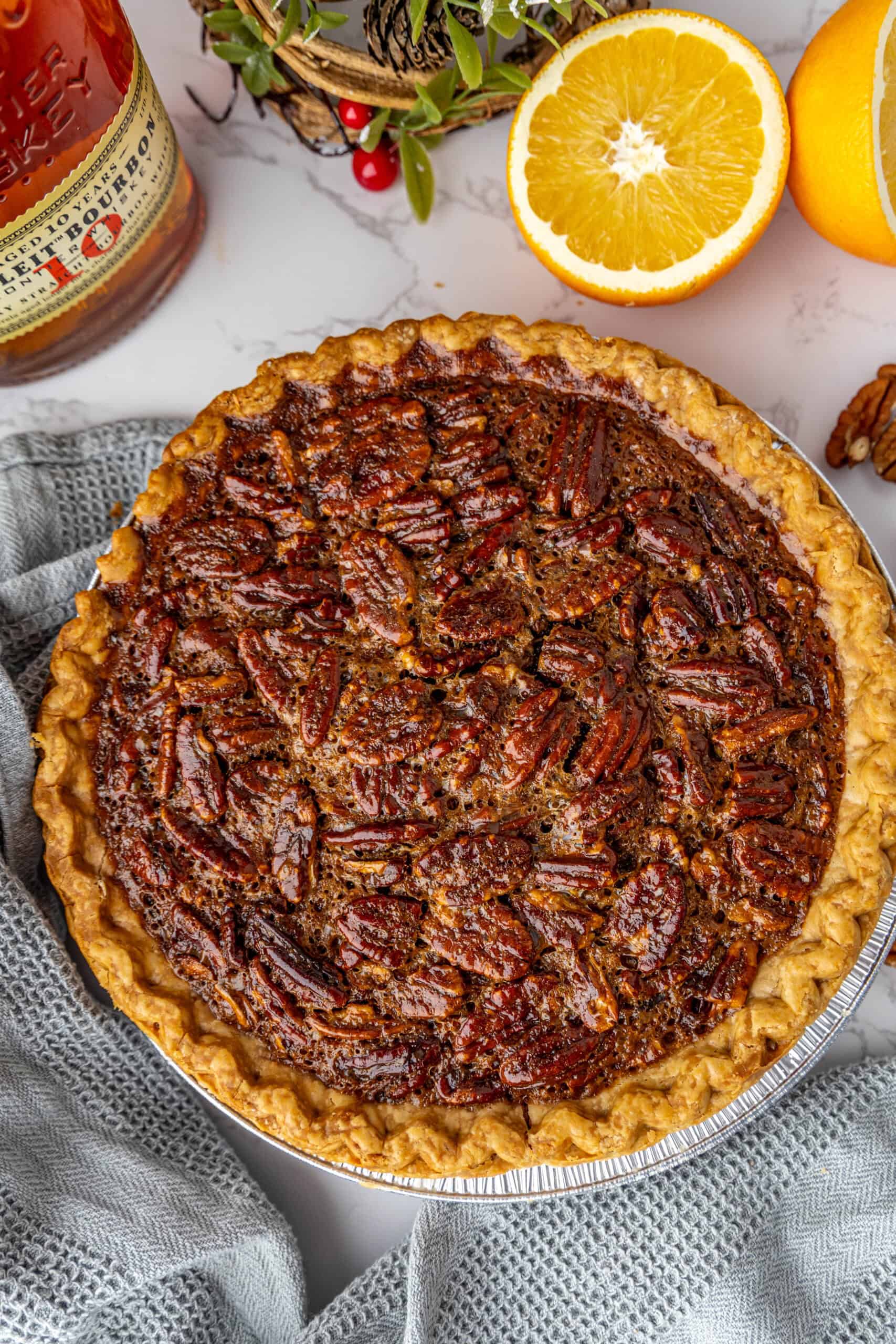 A pecan pie on a table with a halved orange and a bottle in the background, surrounded by a gray cloth.