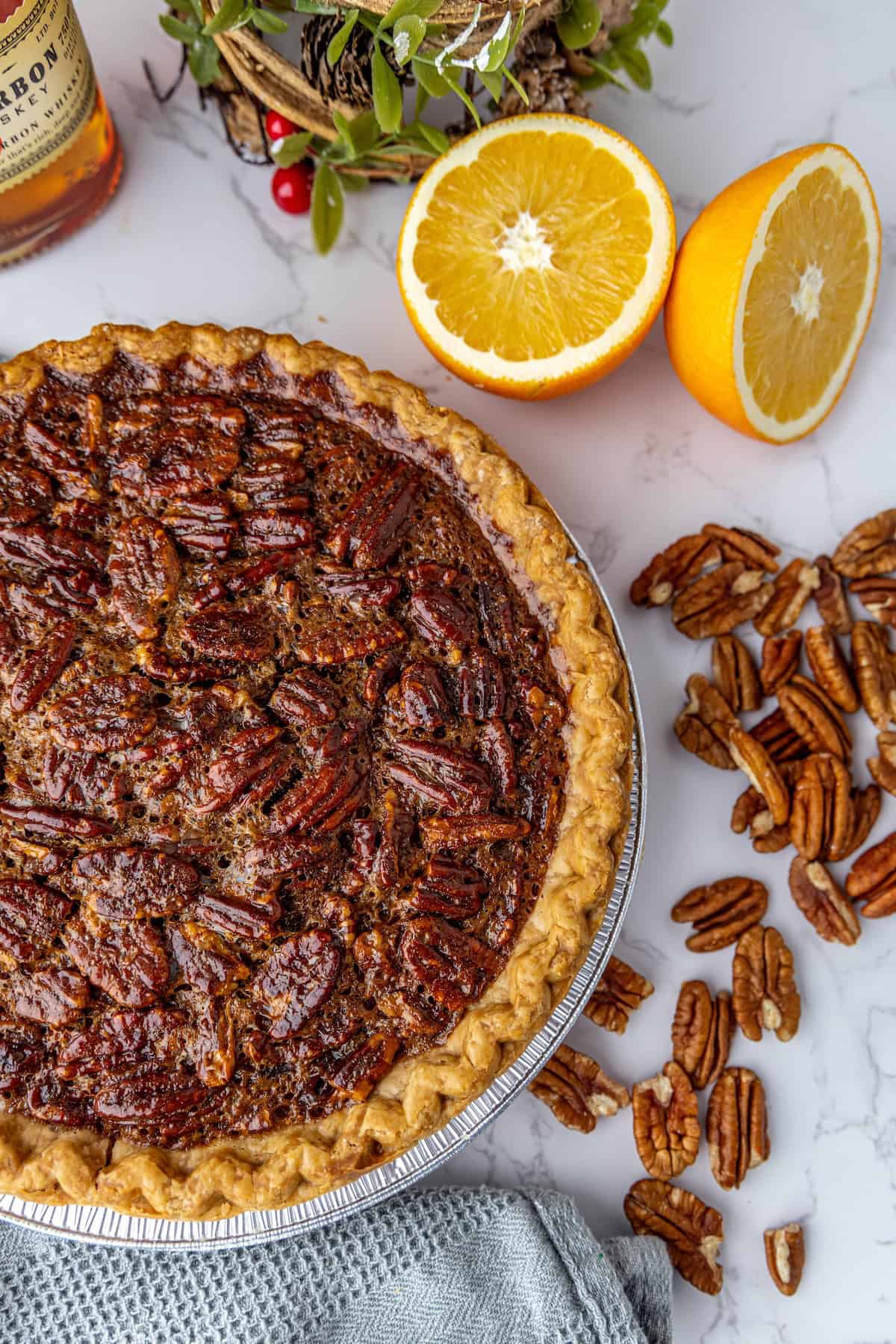 Pecan pie on a marble surface, surrounded by pecans, a halved orange, and a plant.