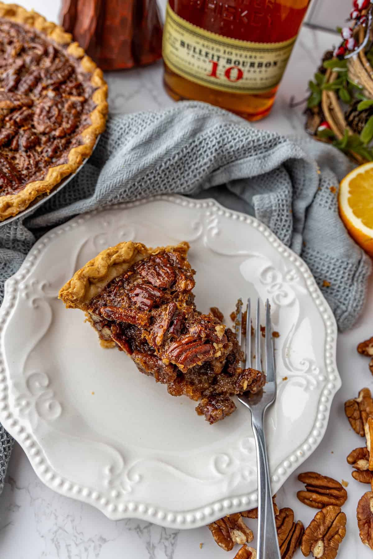 Slice of pecan pie on a white plate with a fork, surrounded by pecans, a bottle of bourbon, and a half orange on a gray cloth background.