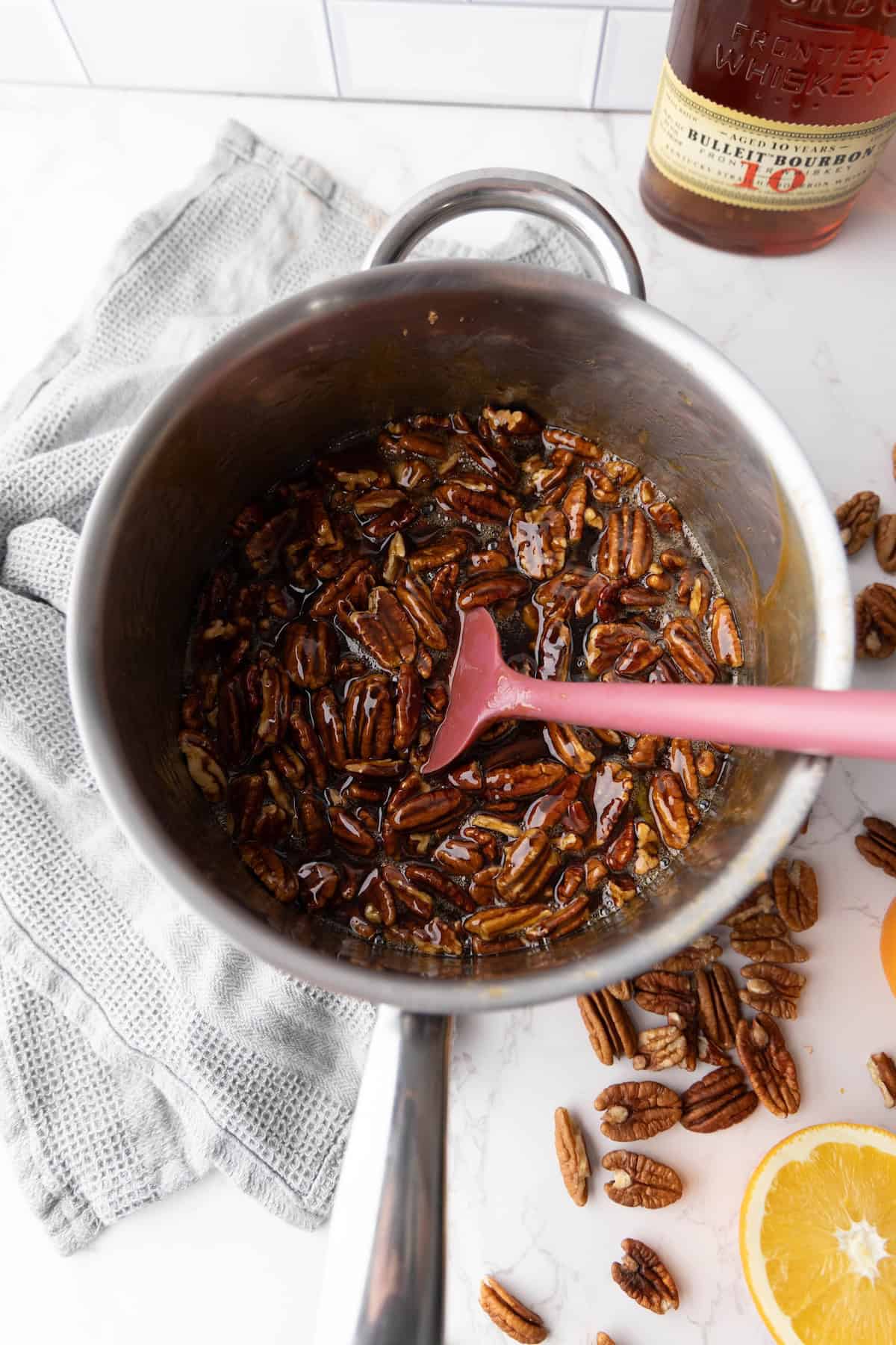 A saucepan of pecans coated in a brown liquid on a marble surface, with a pink spatula inside. A bottle and orange slices are visible in the background.