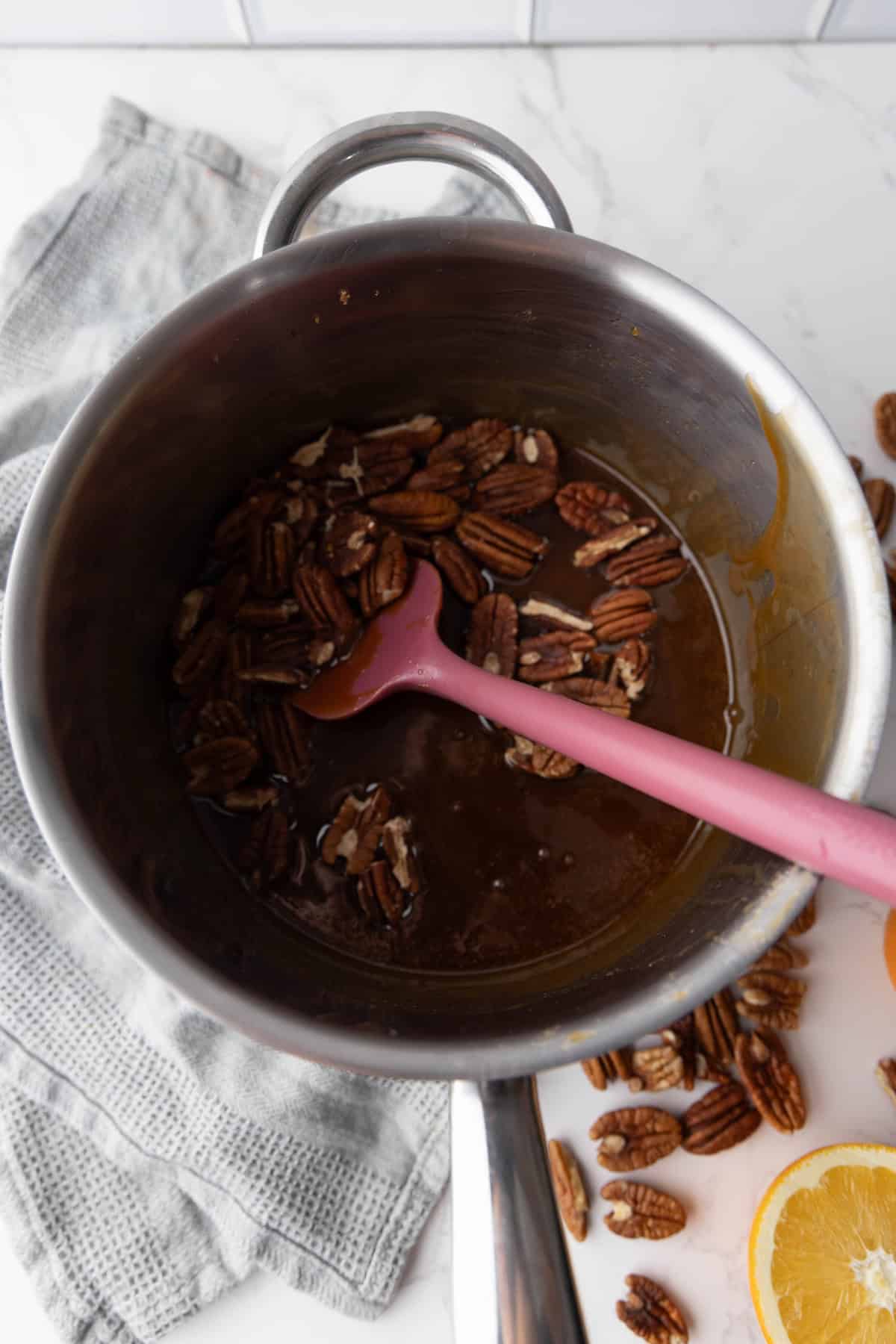 Pot with a spatula stirring pecans in a caramel mixture. Nearby are a gray towel and sliced oranges on a marble surface.