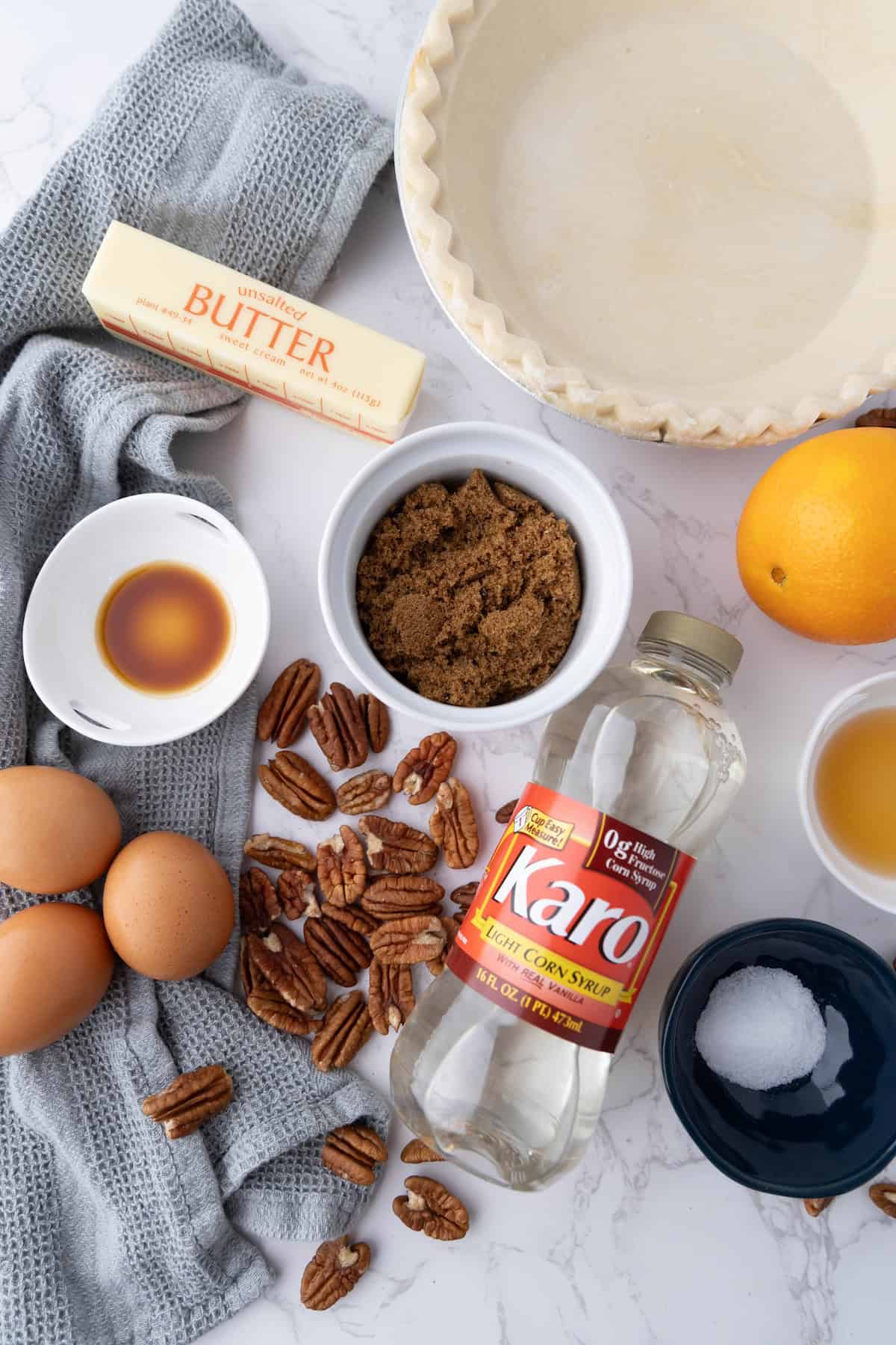 Ingredients for pie preparation, including butter, eggs, brown sugar, light corn syrup, pecans, vanilla extract, an empty pie crust, an orange, and a pinch of salt on a marble surface.