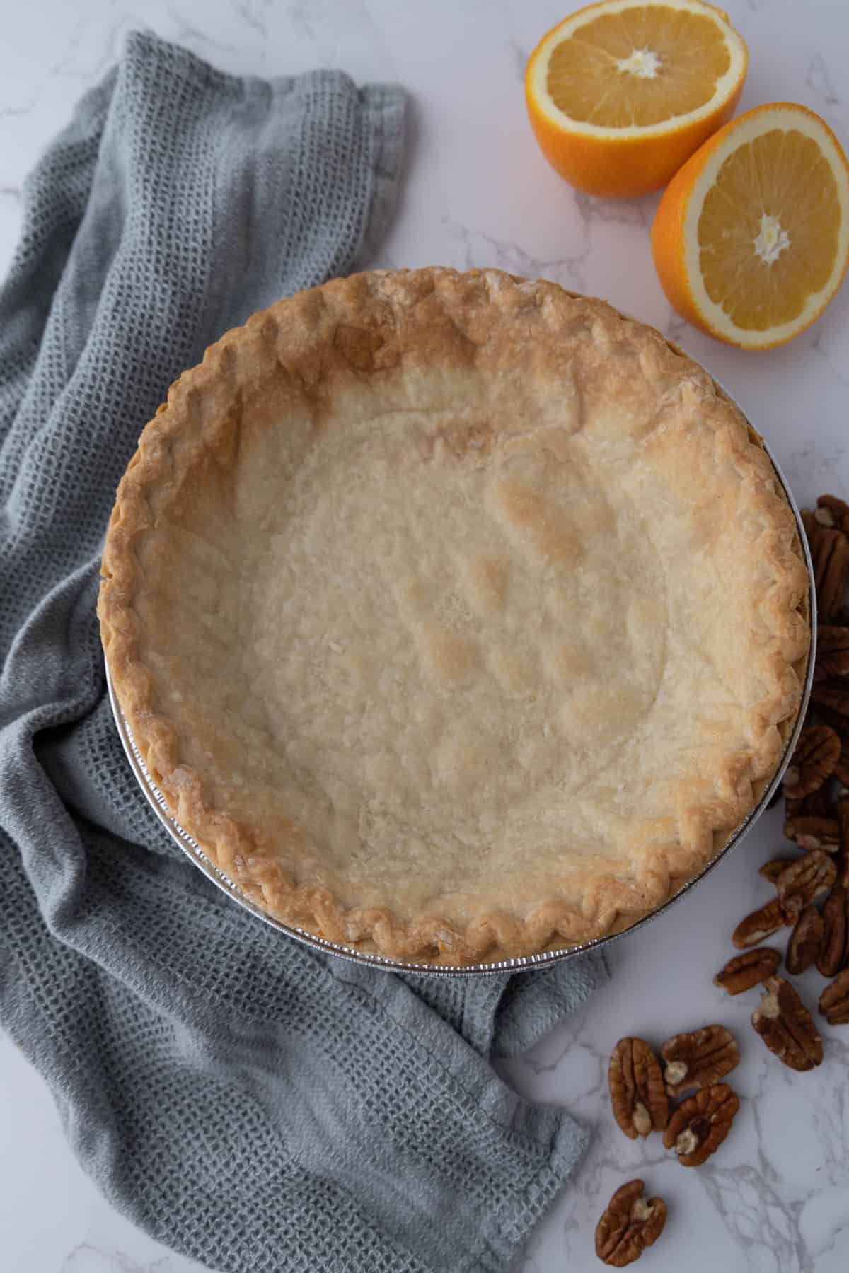 A baked pie crust in a tin next to a gray cloth, halved orange, and scattered pecans on a marble surface.