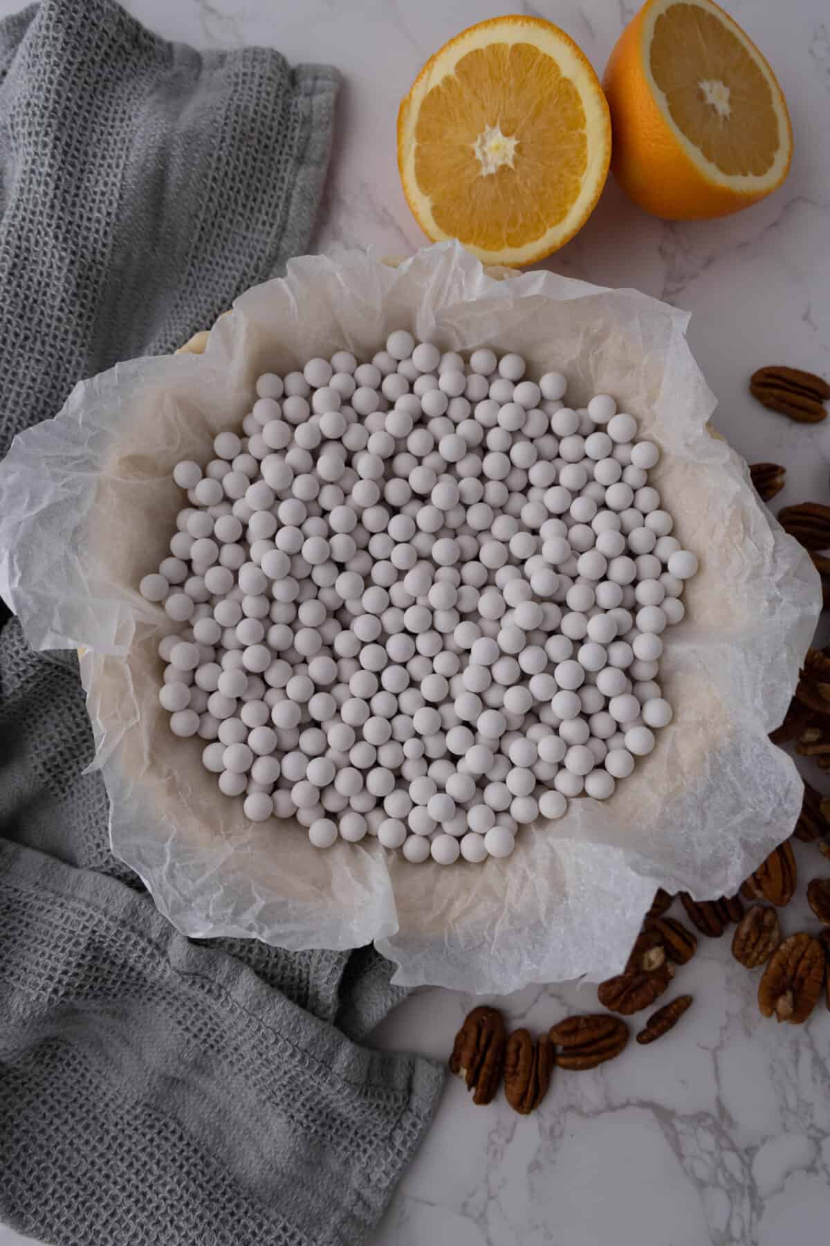 A pie crust lined with parchment paper, filled with white ceramic pie weights, surrounded by pecans and a halved orange on a marble surface.