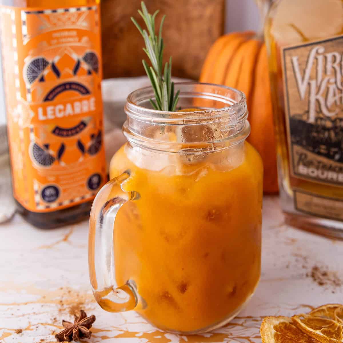 A glass jar filled with an orange-colored cocktail garnished with rosemary. Bottles of alcohol and a small pumpkin are in the background.