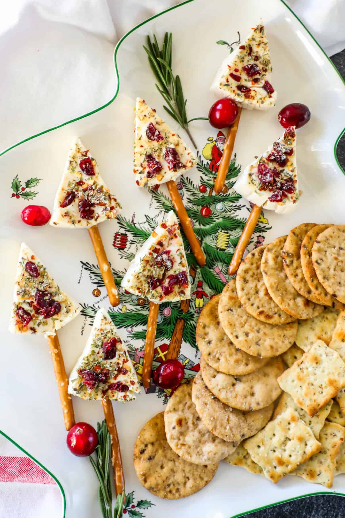 Appetizer plate featuring Christmas tree cheese wedges perched on pretzel sticks, garnished with fresh herbs and cranberries, accompanied by assorted crackers and rosemary sprigs.