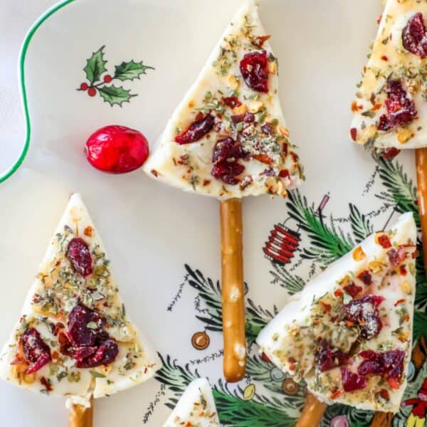 Festive tree-shaped treats and Christmas tree cheese wedges with cranberries and herbs embellish a decorative plate, featuring pretzel sticks as stems and a small red ornament for decoration.