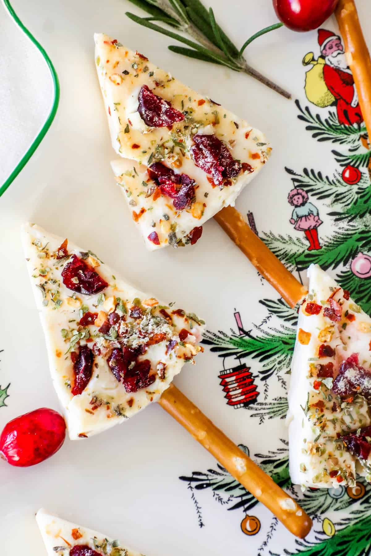 Festive Christmas tree cheese wedges with cranberries and herbs on pretzel sticks, placed on a holiday-themed plate adorned with festive designs and decorations.