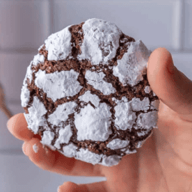 A hand holds one of the delicious chocolate crinkle cookies, its surface artistically cracked and generously dusted with powdered sugar.