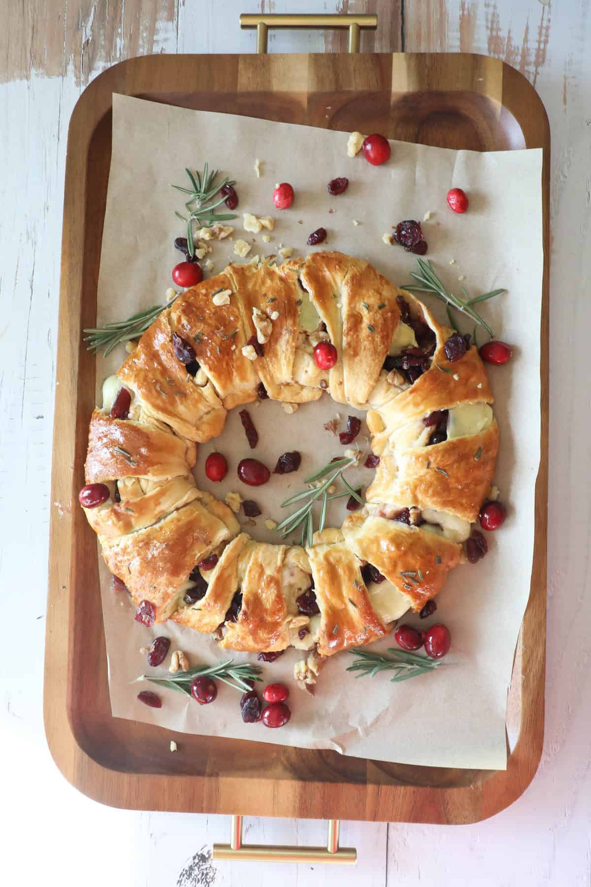 A cranberry brie wreath graces a wooden tray, filled with tangy cranberries and crunchy walnuts, and elegantly garnished with fresh rosemary sprigs.