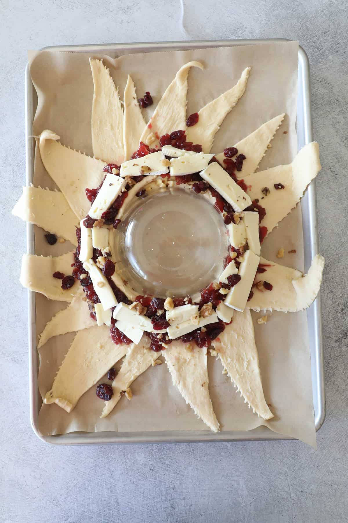 Pastry dough shaped like a sun forms a charming cranberry brie wreath, topped with nuts and dried cranberries, arranged around a glass bowl on a baking sheet.