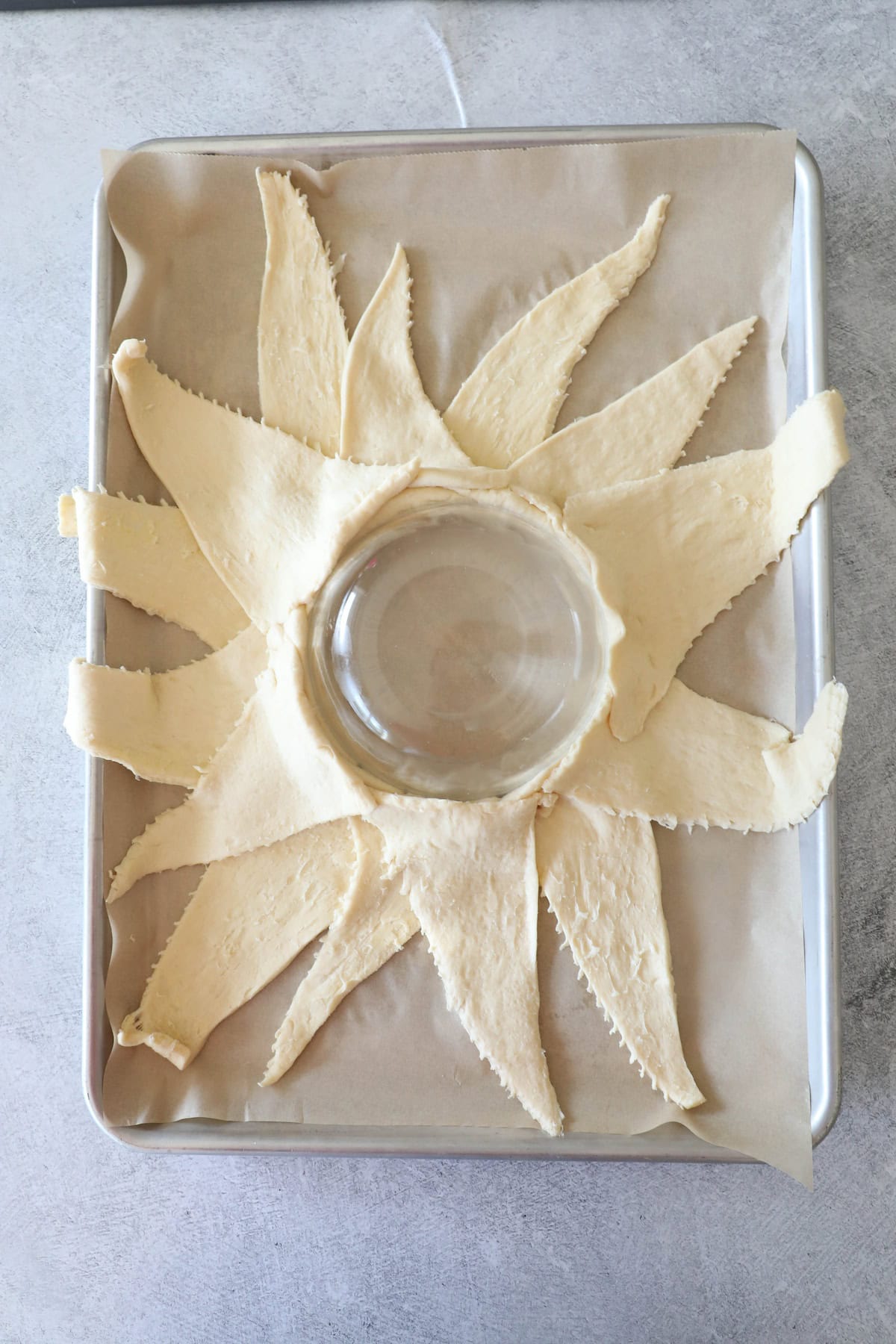 Crescent roll dough pieces are arranged in a starburst pattern on parchment paper with a glass bowl in the center, creating the perfect base for a festive cranberry brie wreath.