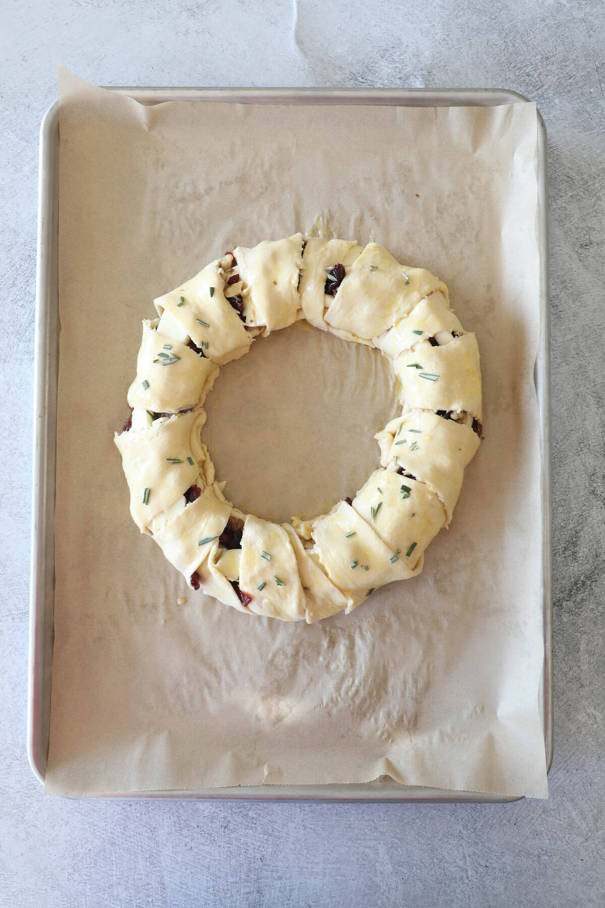 A cranberry brie wreath of unbaked pastry is filled with vegetables and sprinkled with herbs, resting on a parchment-lined baking sheet.