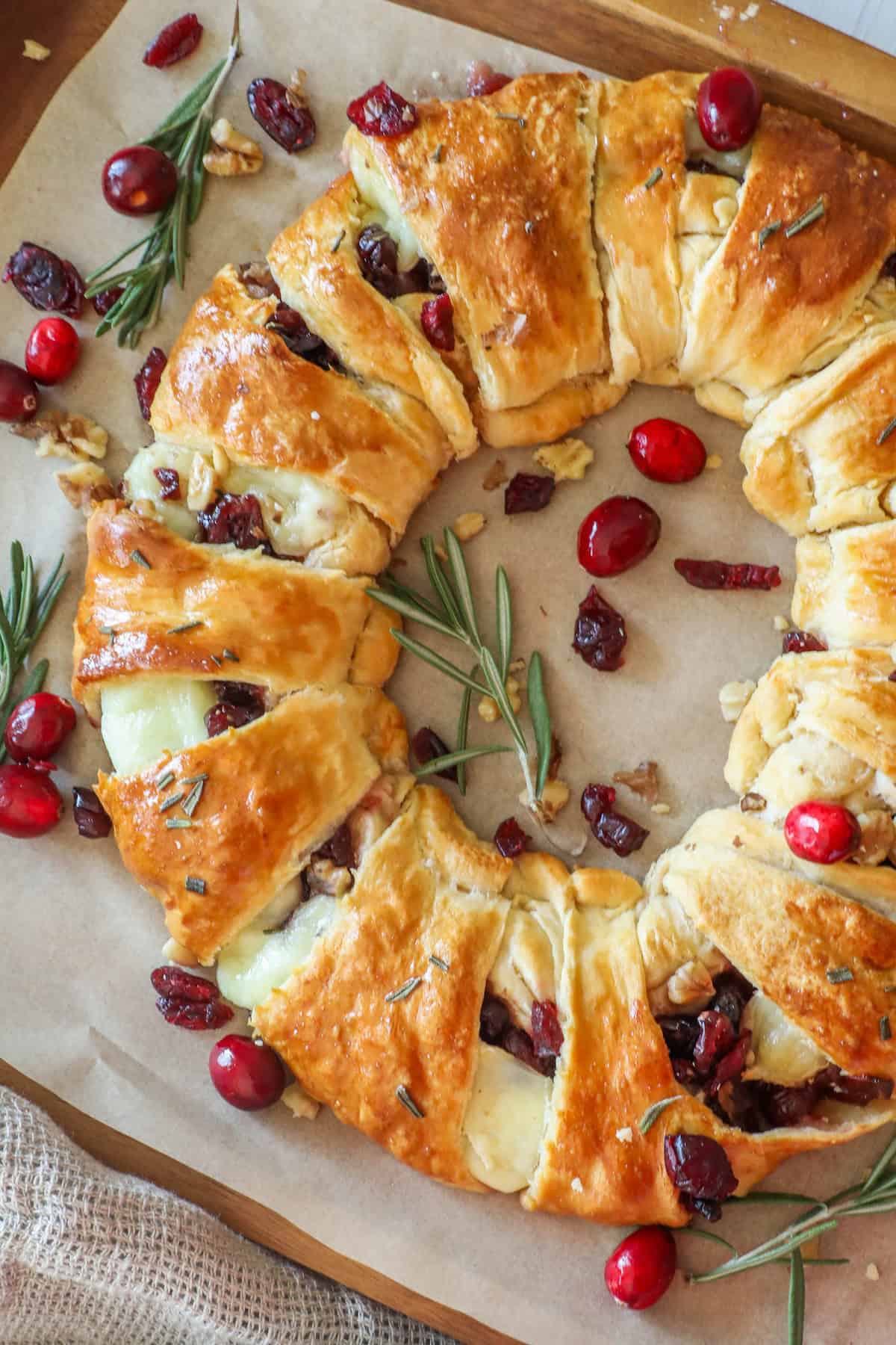 A cranberry brie wreath is a delightful baked pastry wreath featuring melted cheese, cranberries, and rosemary on parchment paper, beautifully garnished with extra cranberries and sprigs of rosemary.