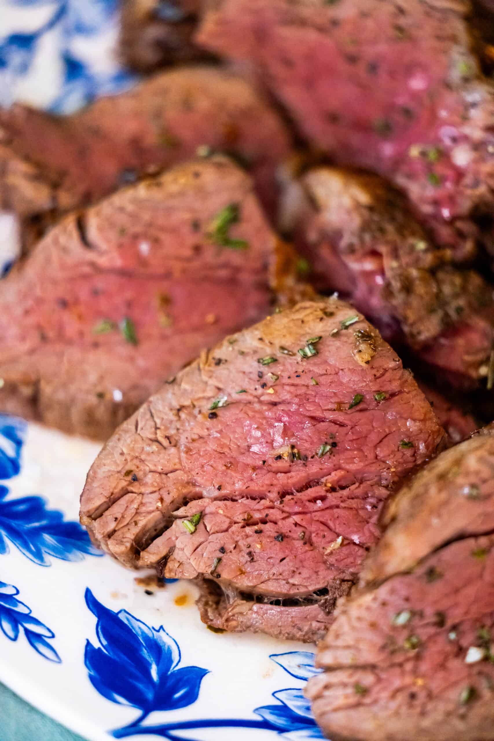 Sliced medium-rare beef tenderloin, prepared using a reverse sear technique, rests elegantly on a blue floral-patterned plate, adorned with fresh herbs.