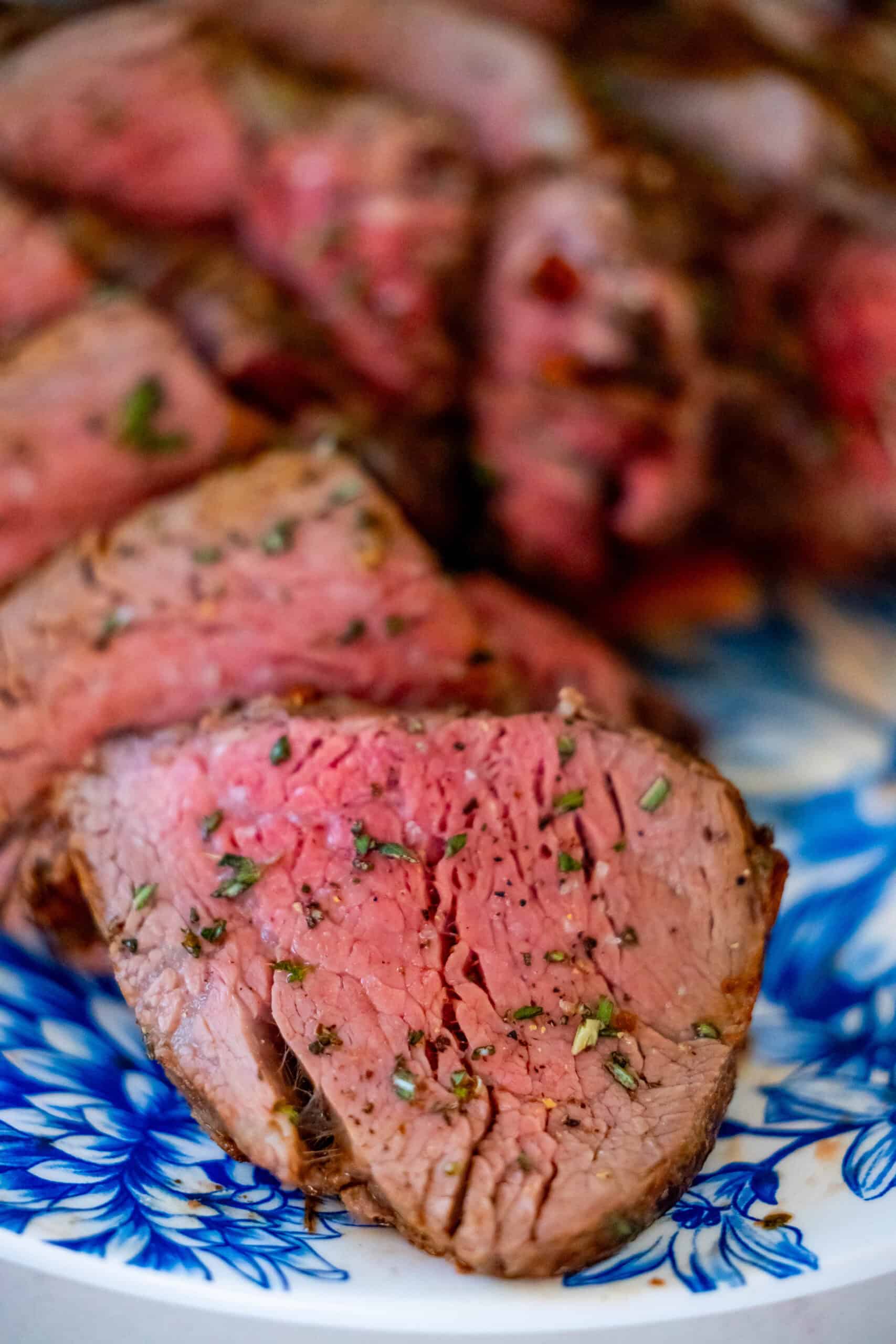 Close-up of sliced medium-rare tenderloin, prepared with a reverse sear technique, garnished with herbs and served on a blue and white patterned plate.